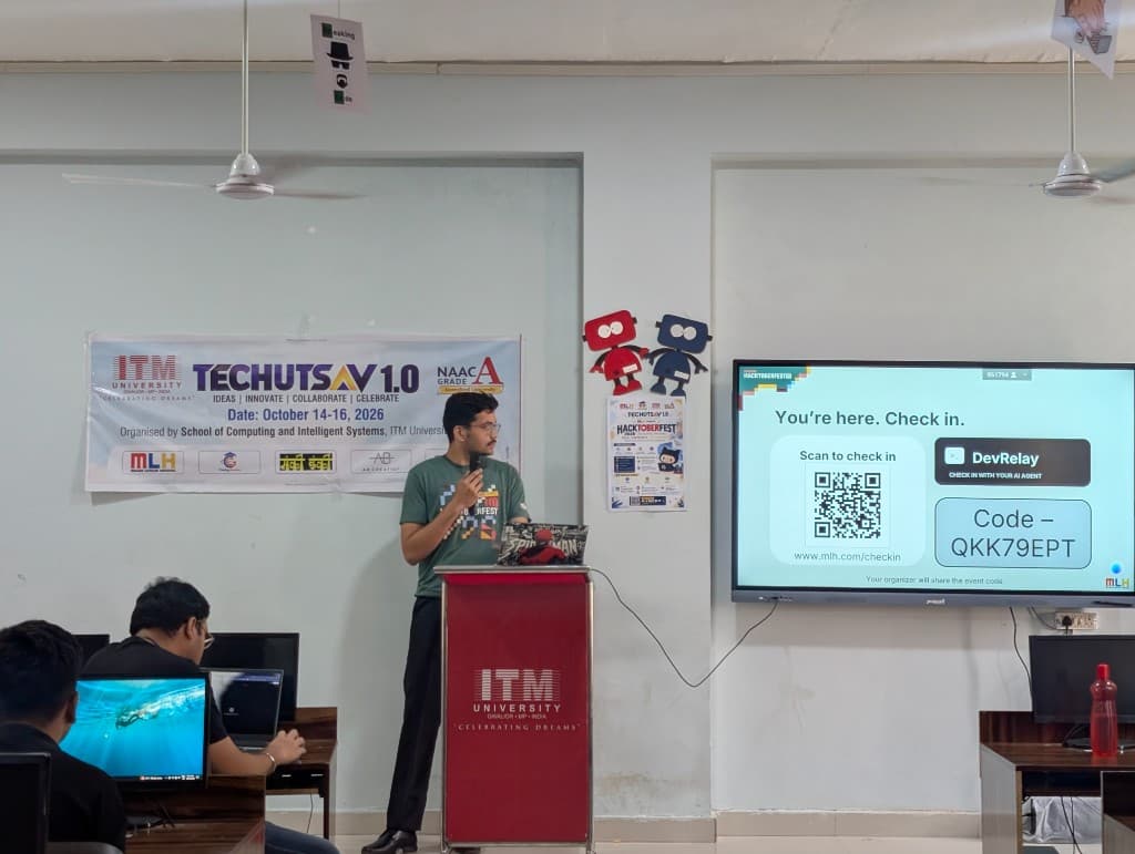 A speaker at the ITM lectern welcoming Hacktoberfest participants, beside a screen showing the MLH check-in code.